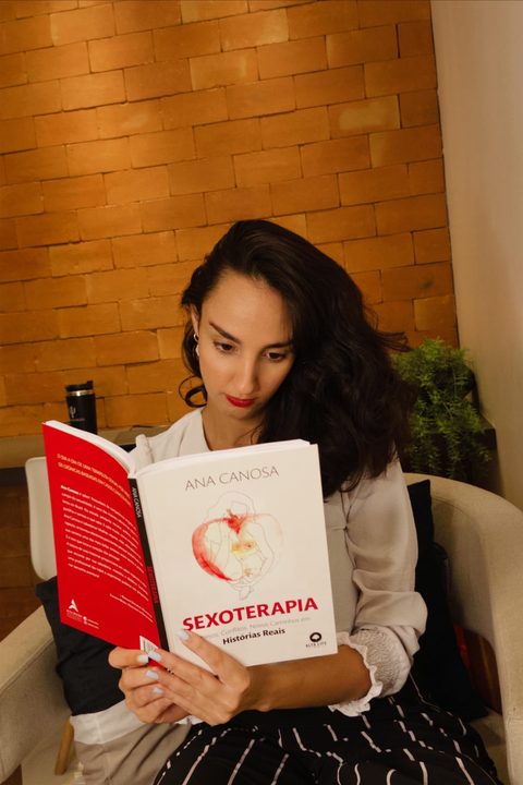 Ana Clara Valoz lendo um livro sobre sexoterapia, sentada numa poltrona