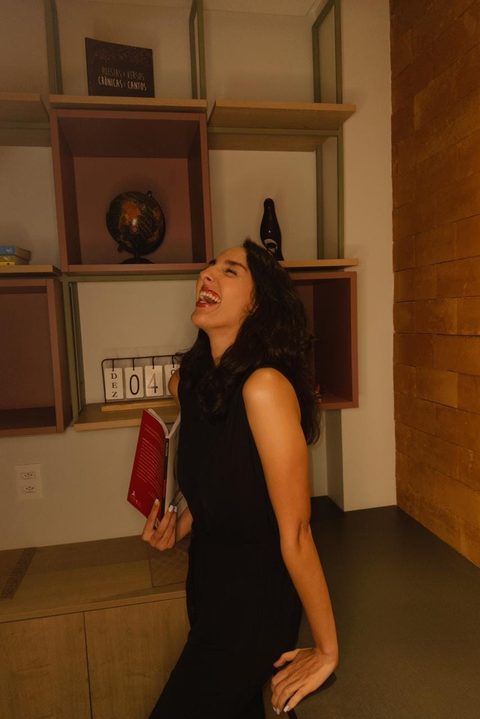 Ana Clara Valoz rindo, com um livro na mão, diante da estante do consultório
